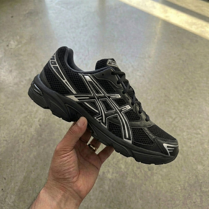 ASICS Gel-1130 Black Pure Silver held in man's hand showing black mesh upper with synthetic leather overlays and metallic silver ASICS stripes against polished concrete background