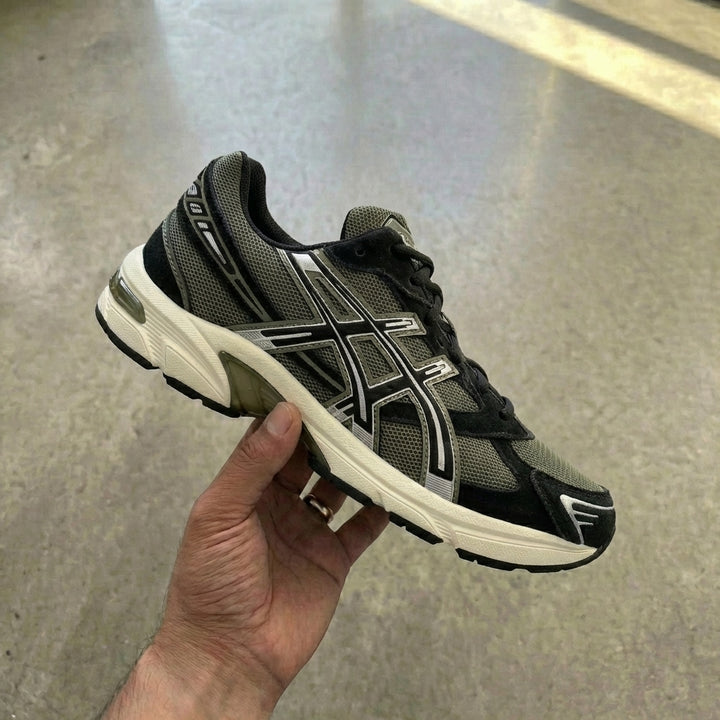 ASICS Gel-1130 Irvine Black held in man's hand showing black mesh upper with Irvine green tones and EVA midsole with GEL insert against polished concrete background