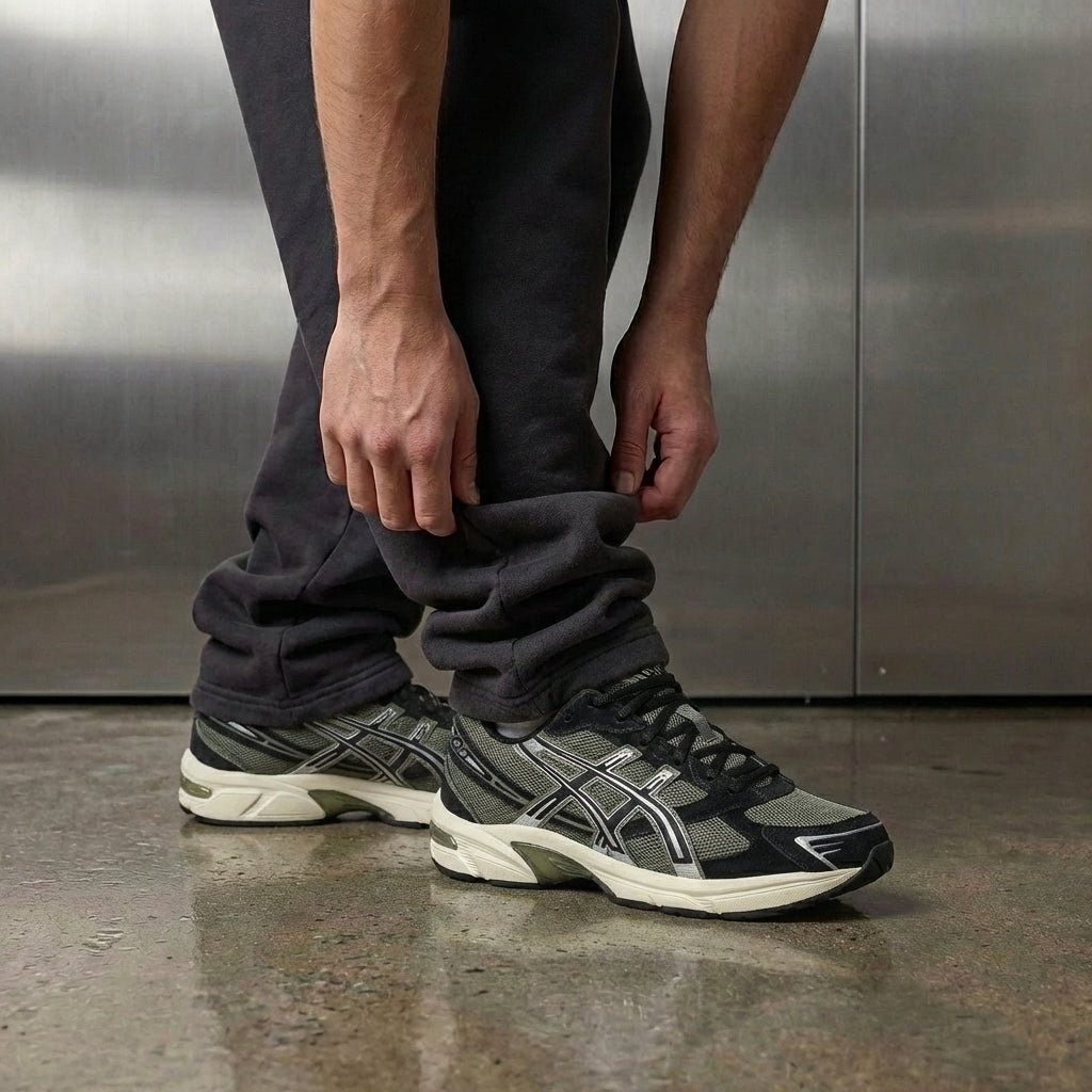 ASICS Gel-1130 Irvine Black on foot, man adjusting jogger cuff to reveal black mesh upper with Irvine green accents and ASICS tiger stripes, industrial studio with stainless steel backdrop and polished concrete floor