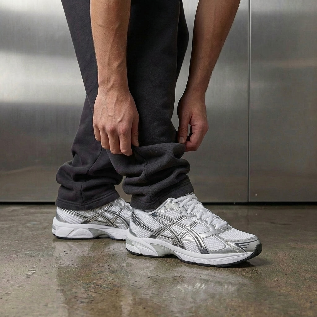 ASICS Gel-1130 White Pure Silver on foot, man adjusting jogger cuff to reveal white mesh upper with pure silver accents and ASICS branding, industrial studio with stainless steel backdrop and polished concrete floor