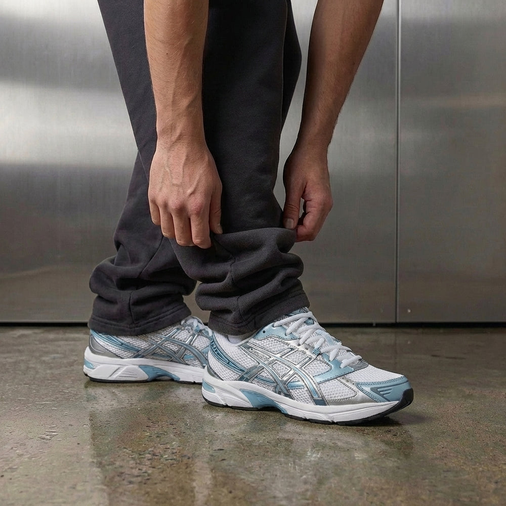 ASICS Gel-1130 White Shark Skin on foot, man adjusting jogger cuff to reveal white mesh upper with steel blue overlays, metallic silver tiger stripes, and visible GEL heel insert, industrial studio with stainless steel backdrop and polished concrete floor