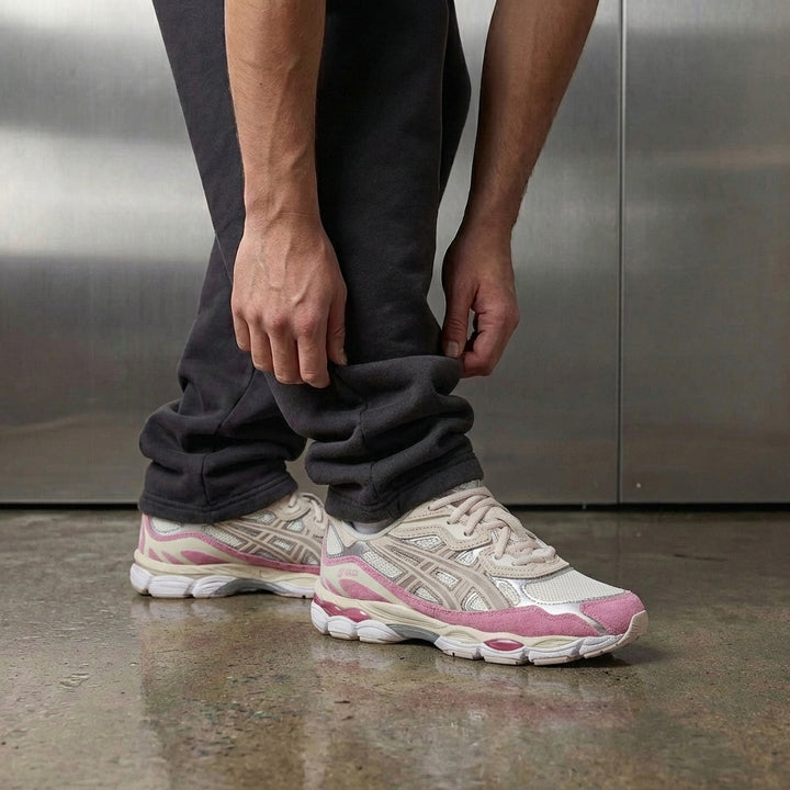 ASICS Gel-NYC Cream Mineral Beige Pink on foot, man adjusting jogger cuff to reveal cream mesh upper with Mineral Beige overlays, pink suede mudguard, and metallic silver accents, industrial studio with stainless steel backdrop and polished concrete floor