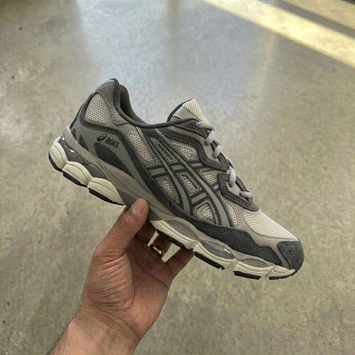 ASICS Gel NYC Oatmeal Obsidian Grey held in man's hand showing oatmeal mesh upper with charcoal suede eyestay, matte silver side stripes, and bicolour foam midsole against polished concrete background