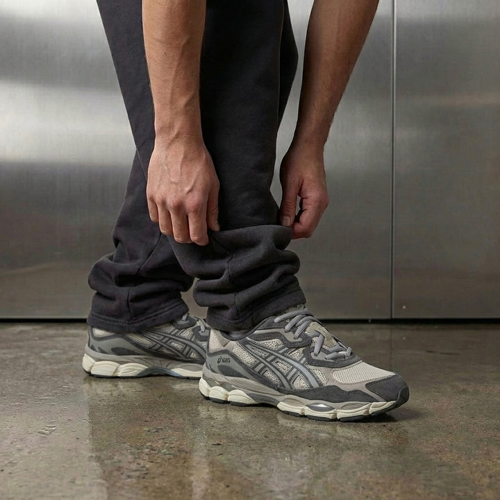 ASICS Gel NYC Oatmeal Obsidian Grey on foot, man adjusting jogger cuff to reveal oatmeal mesh upper with charcoal suede accents and exposed GEL midsole inserts, industrial studio with stainless steel backdrop and polished concrete floor