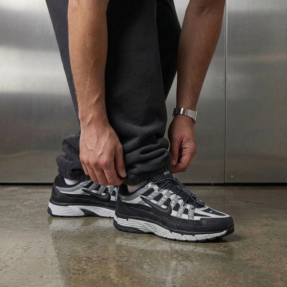 Nike P-6000 Anthracite Smoke Grey on foot, man adjusting jogger cuff to reveal anthracite textile upper with leather overlays and grey foam midsole, industrial studio with stainless steel backdrop and polished concrete floor