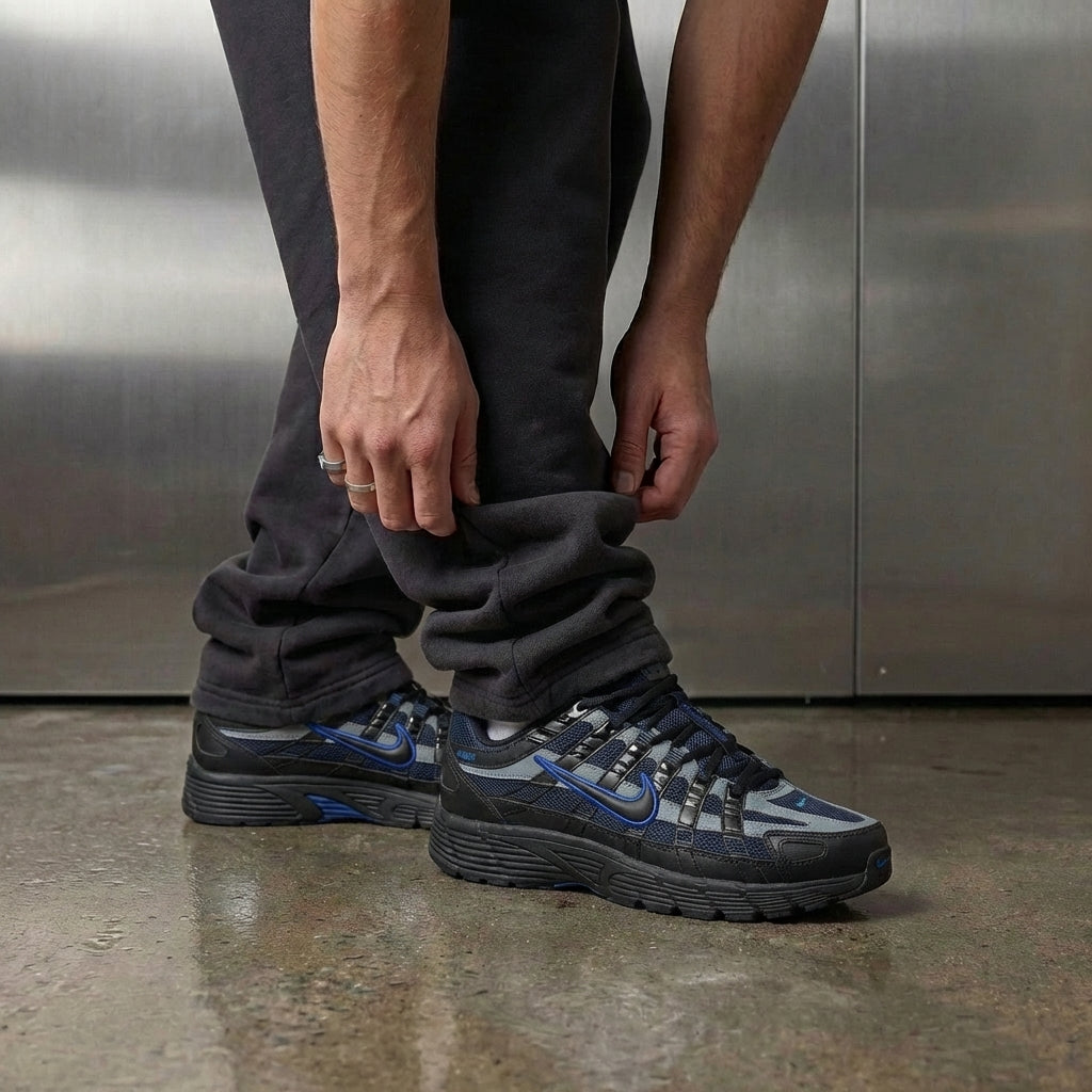 Nike P-6000 Black Obsidian on foot, man adjusting jogger cuff to reveal black mesh upper with obsidian synthetic overlays and cool grey accents, industrial studio with stainless steel backdrop and polished concrete floor