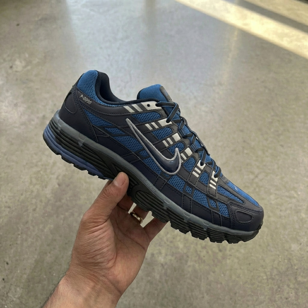 Nike P-6000 Diffused Blue held in man's hand showing black and cool grey mesh upper with synthetic leather overlays and Diffused Blue accents against polished concrete background