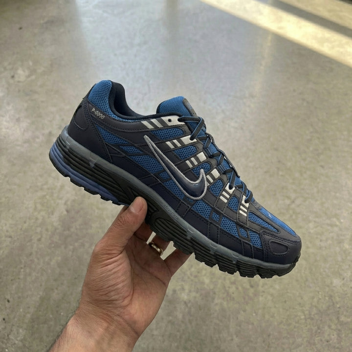 Nike P-6000 Diffused Blue held in man's hand showing black and cool grey mesh upper with synthetic leather overlays and Diffused Blue accents against polished concrete background
