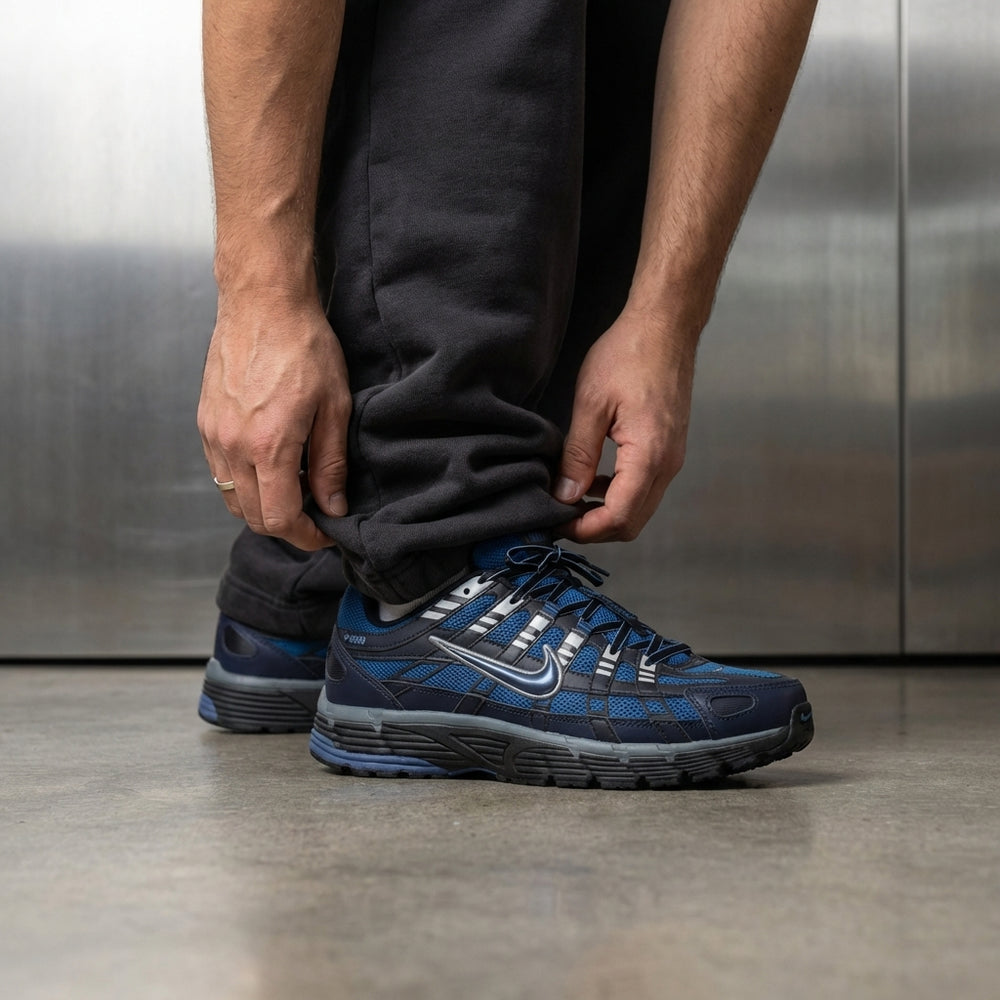 Nike P-6000 Diffused Blue on foot, man adjusting jogger cuff to reveal black and cool grey mesh upper with Diffused Blue Swoosh accents, industrial studio with stainless steel backdrop and polished concrete floor