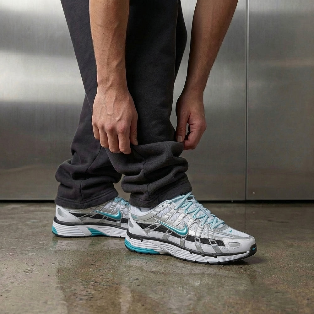 Nike P-6000 Dusty Cactus on foot, man adjusting jogger cuff to reveal white mesh upper with metallic silver and aqua Dusty Cactus leather overlays, industrial studio with stainless steel backdrop and polished concrete floor