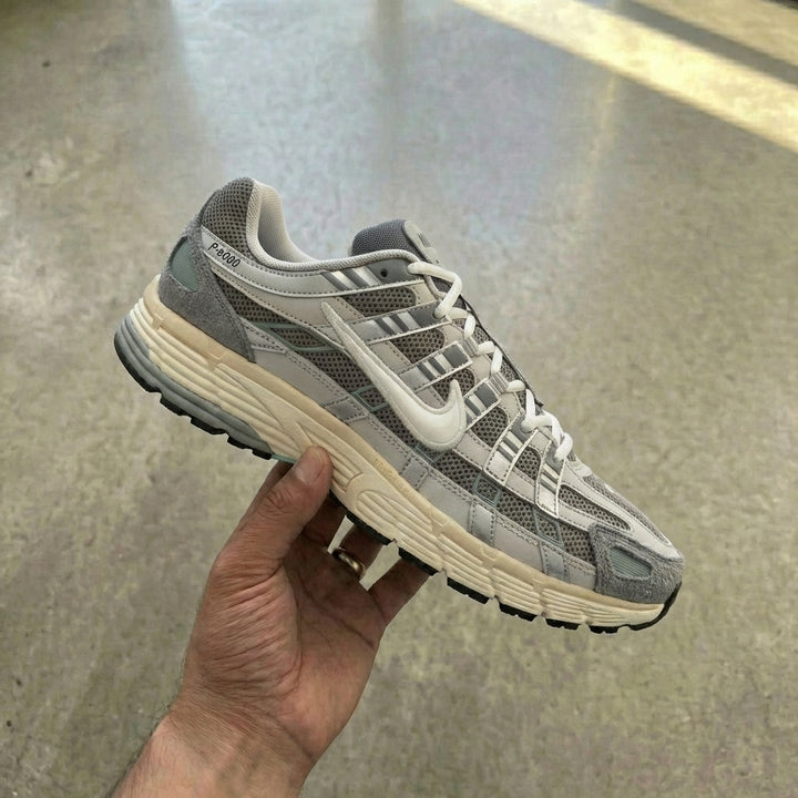 Nike P-6000 Flat Pewter held in man's hand showing matte grey mesh upper with white leather strips, grey suede overlays, and metallic synthetic panels against polished concrete background