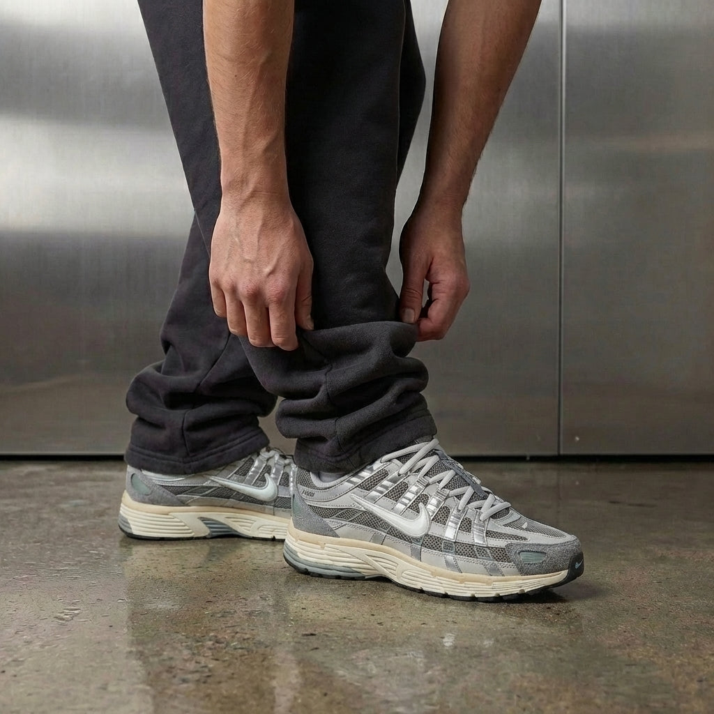 Nike P-6000 Flat Pewter on foot, man adjusting jogger cuff to reveal matte grey mesh upper with suede toe, white leather Swoosh, and aqua-green TPU accents, industrial studio with stainless steel backdrop and polished concrete floor