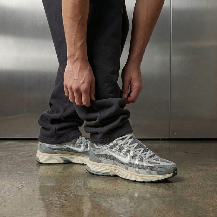 Nike P-6000 Flat Pewter on foot, man adjusting jogger cuff to reveal matte grey mesh upper with suede toe, white leather Swoosh, and aqua-green TPU accents, industrial studio with stainless steel backdrop and polished concrete floor