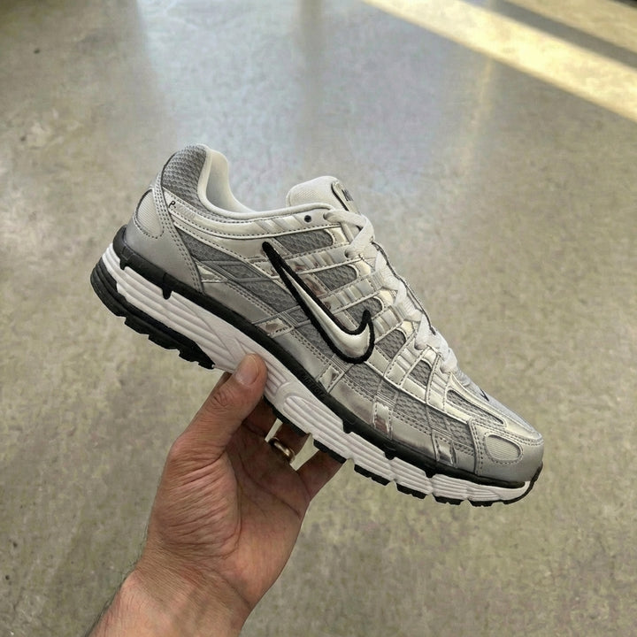 Nike P-6000 Metallic Silver held in man's hand showing chrome silver mesh upper with shiny synthetic leather overlays and floating Swoosh against polished concrete background