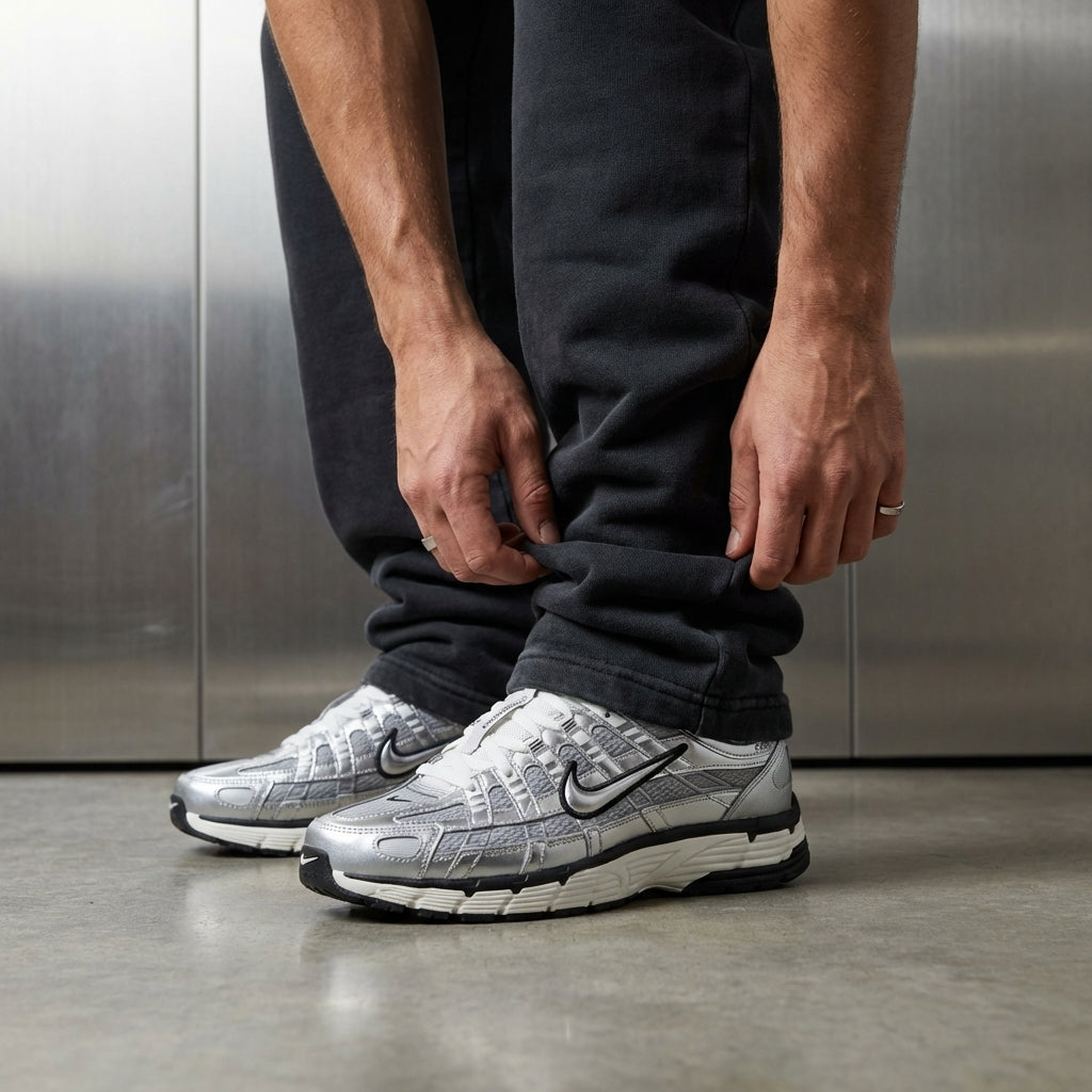 Nike P-6000 Metallic Silver on foot, man adjusting jogger cuff to reveal chrome silver mesh upper with synthetic leather overlays and black embroidered Swoosh, industrial studio with stainless steel backdrop and polished concrete floor