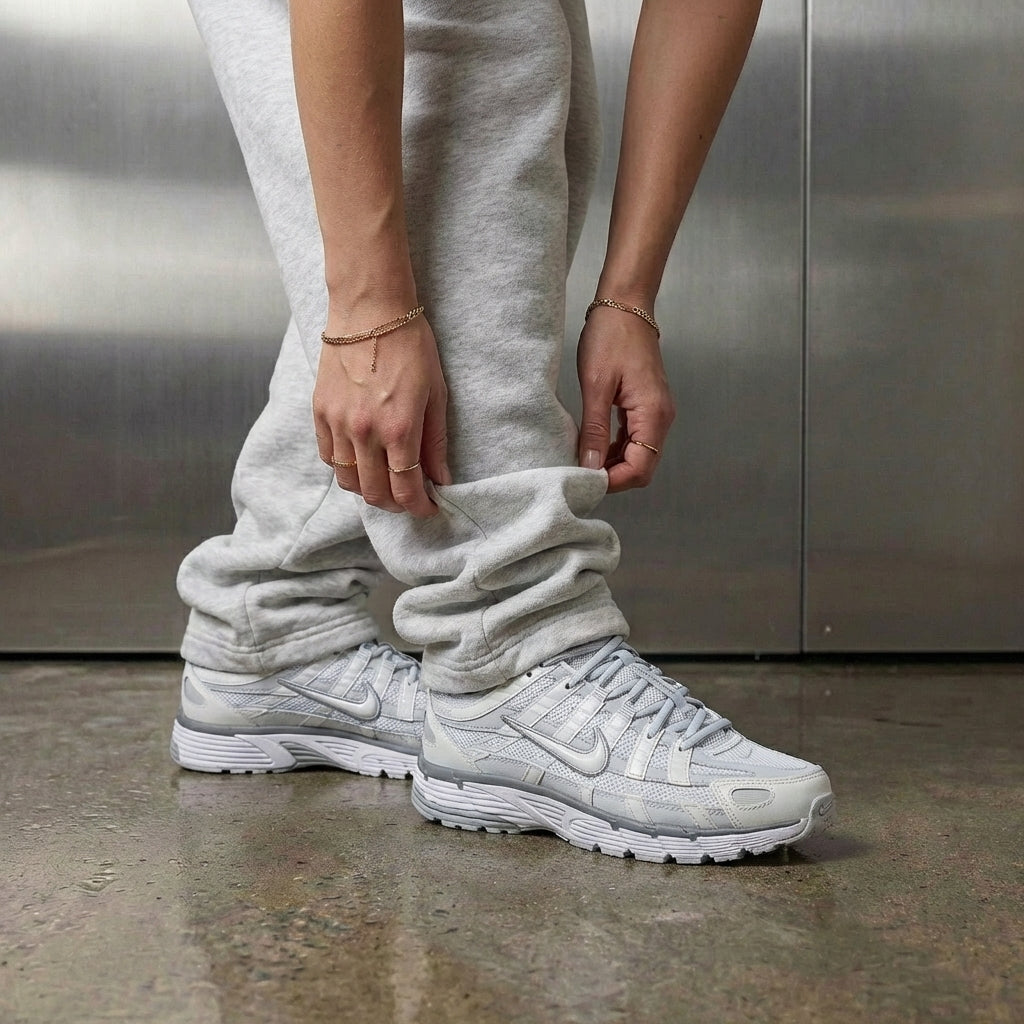 Nike P-6000 Summit White Pure Platinum on foot, woman adjusting grey jogger cuff to reveal summit white mesh upper with pure platinum overlays, industrial studio with stainless steel backdrop and polished concrete floor