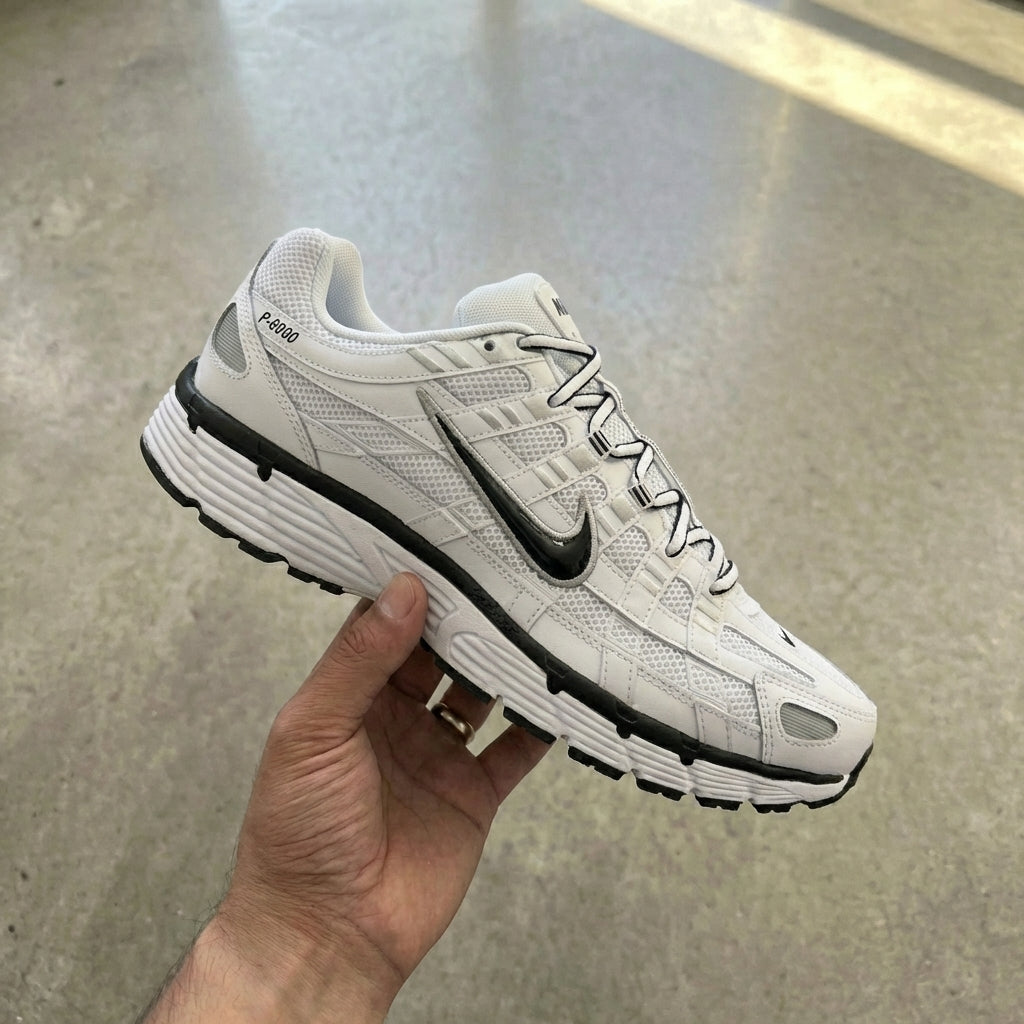 Nike P-6000 White Metallic Silver Black held in man's hand showing white mesh upper with leather overlays and reflective metallic silver detailing against polished concrete background