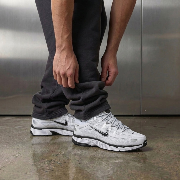 Nike P-6000 White Metallic Silver Black on foot, man adjusting jogger cuff to reveal white mesh upper with metallic silver reflective details and black Swoosh accents, industrial studio with stainless steel backdrop and polished concrete floor