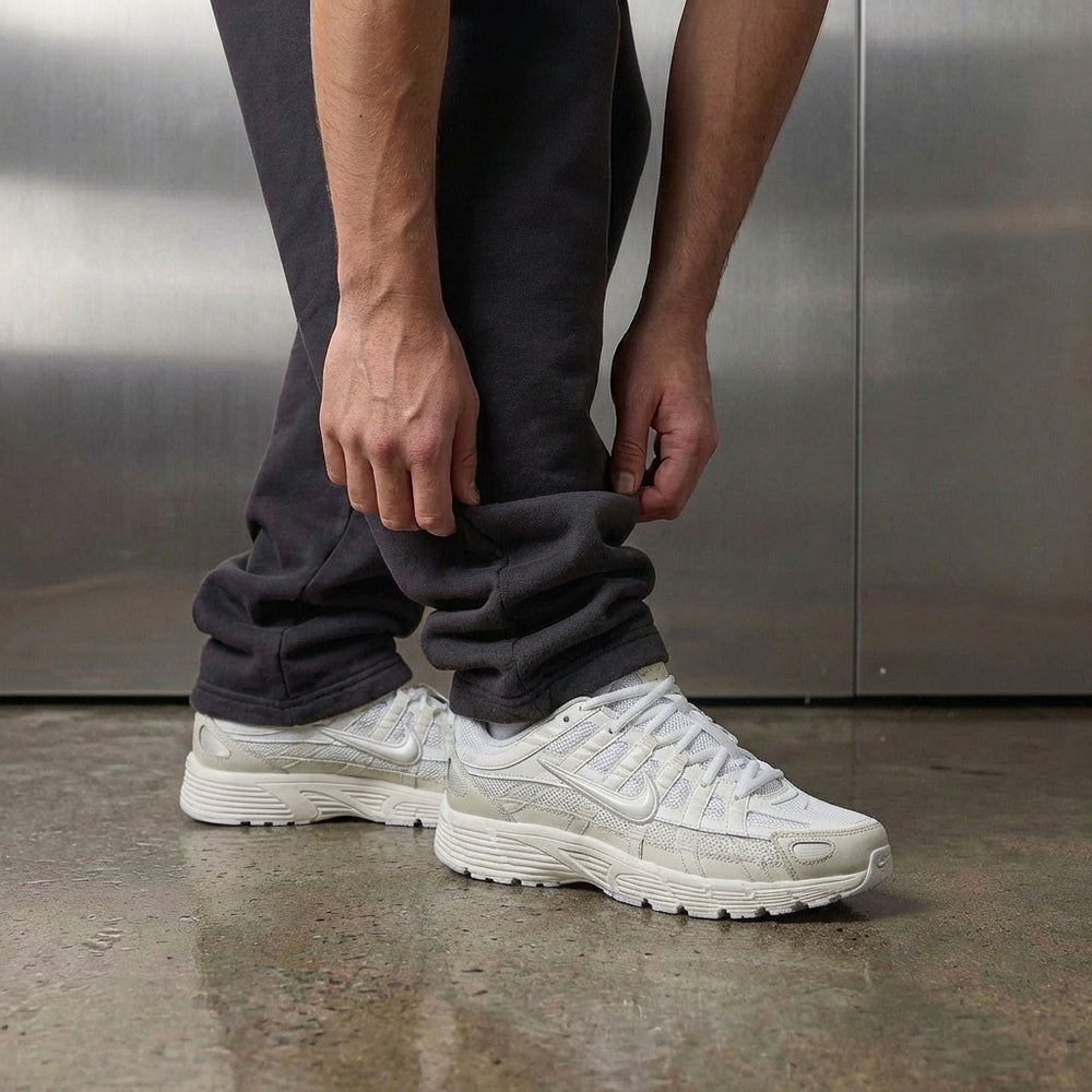 Nike P-6000 White Sail on foot, man adjusting jogger cuff to reveal white mesh upper with sail cream and vast grey overlays, industrial studio with stainless steel backdrop and polished concrete floor