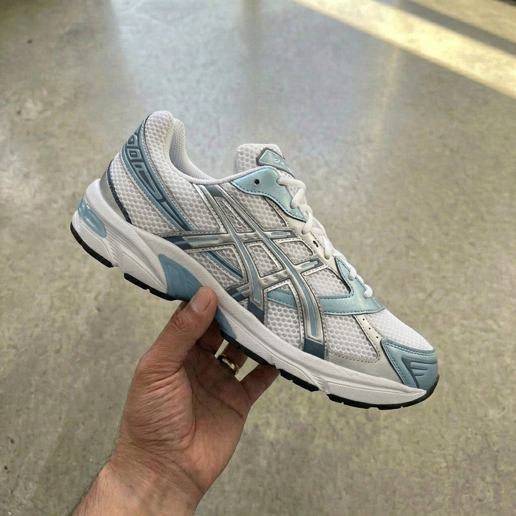 ASICS Gel-1130 White Shark Skin held in man's hand showing white mesh upper with steel blue synthetic leather overlays and metallic silver tiger stripes against polished concrete background