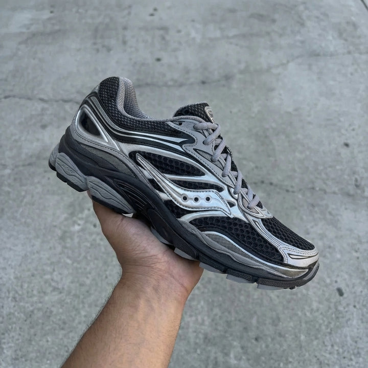 Saucony ProGrid Omni 9 Silver Black held in man's hand showing black and silver synthetic and leather upper with reflective detailing against polished concrete background
