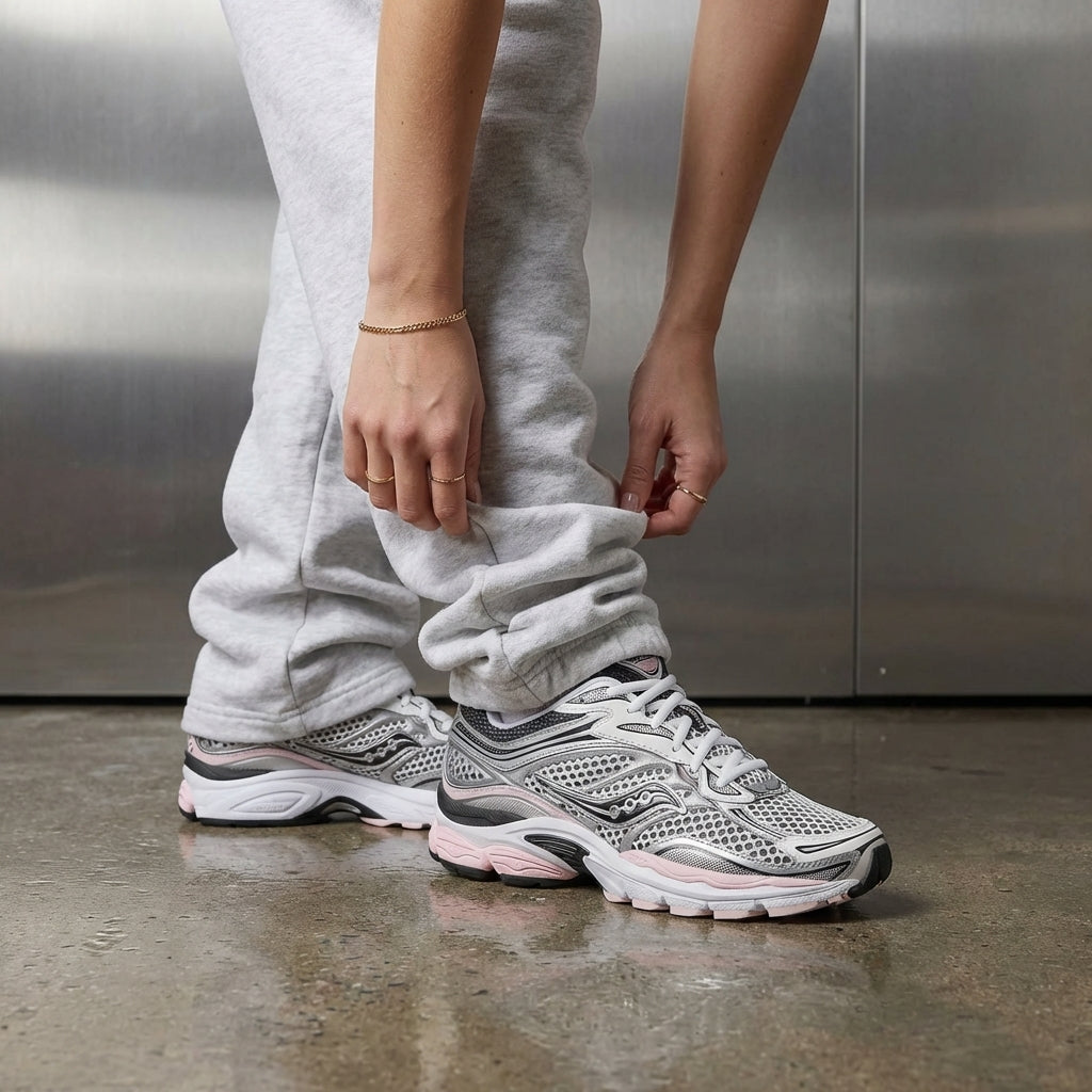 Saucony ProGrid Omni 9 Silver Pink on foot, woman adjusting grey jogger cuff to reveal silver mesh upper and pastel pink midsole, industrial studio with stainless steel backdrop and polished concrete floor